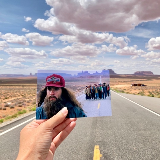 Hand is holding a still photo of man with group of people behind him on a road. This matches up with the same position on the road the person is standing with red rock formations in the background.